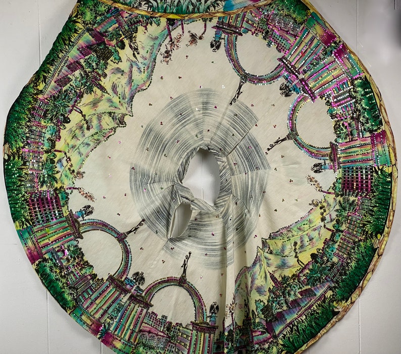 May include: A white fabric circle with a colorful, detailed design of a city skyline and arches. The fabric is decorated with small, iridescent sequins.
