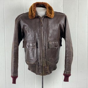 Puede incluir: Una chaqueta bomber de cuero marrón con cuello de piel de oveja y cierre de cremallera. La chaqueta tiene dos bolsillos de solapa en la parte delantera y puños de punto de canalé.