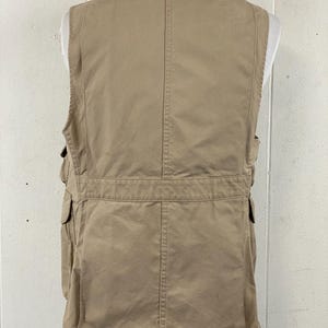 May include: Tan utility vest with multiple pockets and a high collar. The vest is displayed on a mannequin and has a classic design. The vest is made of a durable material and is suitable for outdoor activities.