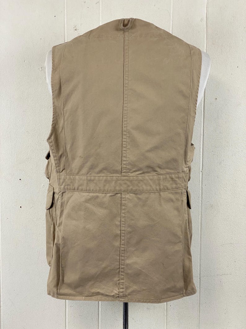 May include: Tan utility vest with multiple pockets and a high collar. The vest is displayed on a mannequin and has a classic design. The vest is made of a durable material and is suitable for outdoor activities.
