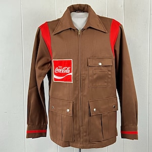 Pode incluir: Um casaco castanho com fecho de correr, painéis vermelhos brilhantes nos ombros e punhos. A frente apresenta um emblema retangular com o texto branco Enjoy Coca-Cola num fundo vermelho. Inclui dois bolsos no peito e dois bolsos inferiores.