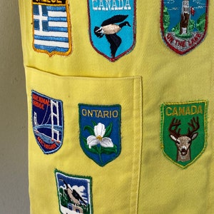 May include: Yellow fabric vest with various embroidered patches, including "Greece", "Canada", "Niagara on the Lake", "Mackinac Bridge", "Ontario", "Wawa", and "Canada" with a deer.