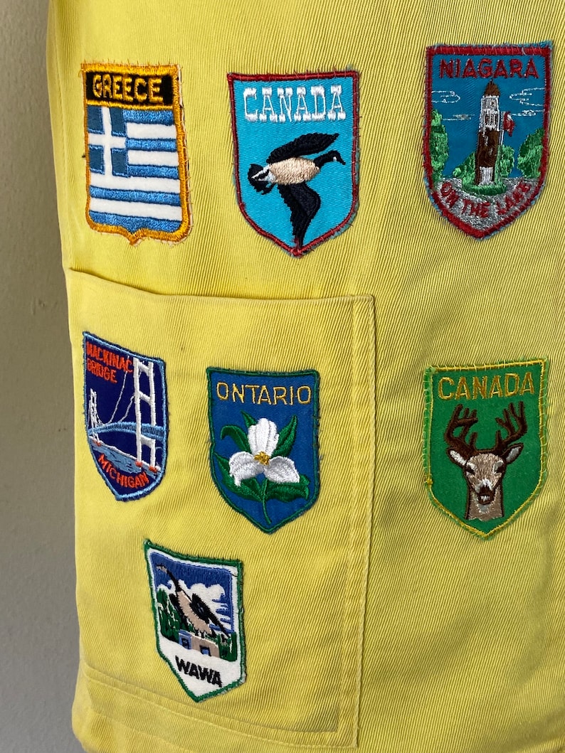 May include: Yellow fabric vest with various embroidered patches, including "Greece", "Canada", "Niagara on the Lake", "Mackinac Bridge", "Ontario", "Wawa", and "Canada" with a deer.