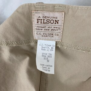 May include: Close-up of a tan garment with a Filson brand label. The label reads "A Genuine Filson" and includes the text "Might as well have the best." A smaller tag indicates the garment's style, size S, and material: 100% cotton.