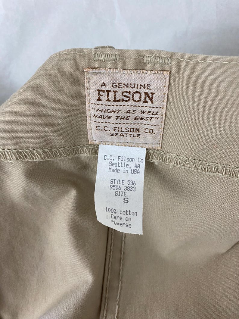 May include: Close-up of a tan garment with a Filson brand label. The label reads "A Genuine Filson" and includes the text "Might as well have the best." A smaller tag indicates the garment's style, size S, and material: 100% cotton.