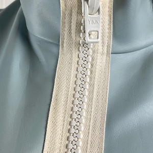 K&ouml;nnte beinhalten: Nahaufnahme eines wei&szlig;en Rei&szlig;verschlusses mit einem YKK-Logo auf einer hellblauen Jacke.