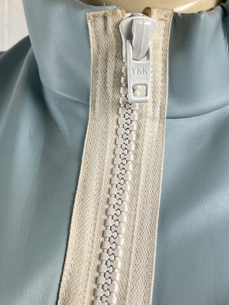 K&ouml;nnte beinhalten: Nahaufnahme eines wei&szlig;en Rei&szlig;verschlusses mit einem YKK-Logo auf einer hellblauen Jacke.