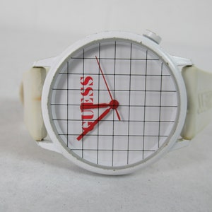 May include: White wristwatch with a white strap and a white face. The face has a grid pattern and the word "GUESS" in red letters.