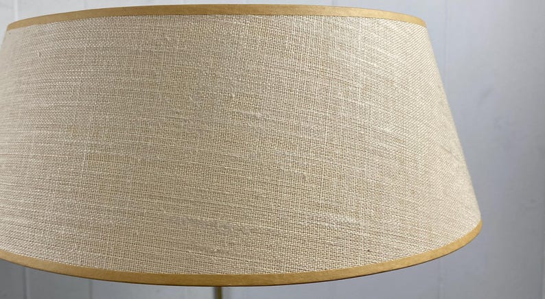 May include: A beige lampshade with a textured fabric. The lampshade has a tapered shape and is trimmed with a thin, gold-coloured band at the top and bottom. The neutral colour and simple design suggest a classic style.