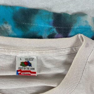 Puede incluir: Una camiseta blanca Fruit of the Loom con una etiqueta de talla L. La etiqueta es blanca con acentos rojos, azules y verdes. La etiqueta dice "100% algod&oacute;n preencogido" y "Fruit of the Loom" con "Hecho en EE. UU." debajo.