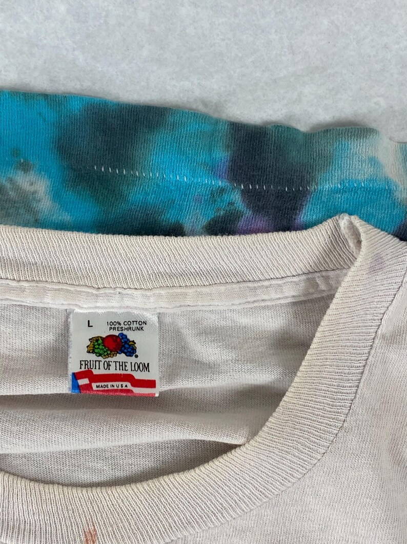 Puede incluir: Una camiseta blanca Fruit of the Loom con una etiqueta de talla L. La etiqueta es blanca con acentos rojos, azules y verdes. La etiqueta dice "100% algod&oacute;n preencogido" y "Fruit of the Loom" con "Hecho en EE. UU." debajo.