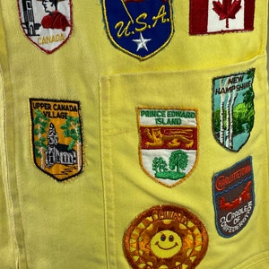May include: Yellow fabric with various embroidered patches, including a smiling sun, a Canadian flag, a US flag, and patches with text like "Ottawa, Canada", "Upper Canada Village", "Prince Edward Island", "New Hampshire", and "Charlottetown, Cradle of Confederation".