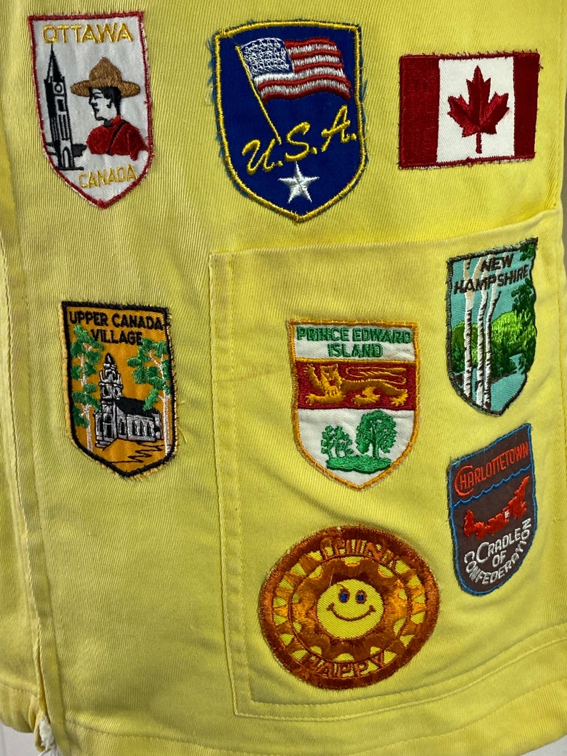 May include: Yellow fabric with various embroidered patches, including a smiling sun, a Canadian flag, a US flag, and patches with text like "Ottawa, Canada", "Upper Canada Village", "Prince Edward Island", "New Hampshire", and "Charlottetown, Cradle of Confederation".