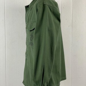 May include: Olive green button-up shirt with two chest pockets. The shirt has a collared neckline and long sleeves with buttoned cuffs. The fabric appears to be a durable cotton or blend. The shirt has a military-inspired design.