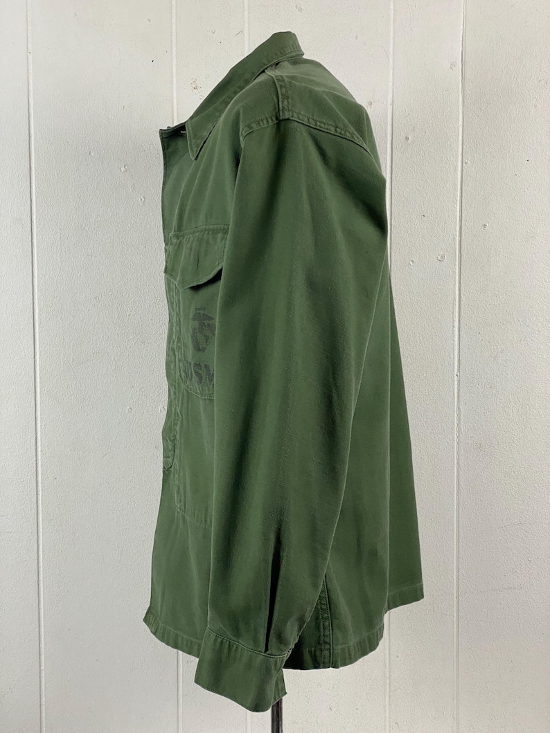 May include: Olive green button-up shirt with two chest pockets. The shirt has a collared neckline and long sleeves with buttoned cuffs. The fabric appears to be a durable cotton or blend. The shirt has a military-inspired design.