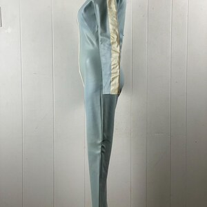 K&ouml;nnte beinhalten: Ein hellblauer Jumpsuit mit einem wei&szlig;en Streifen an der Seite des rechten Beins und &Auml;rmels. Der Jumpsuit hat einen Rei&szlig;verschluss an der Vorderseite und einen hohen Kragen.
