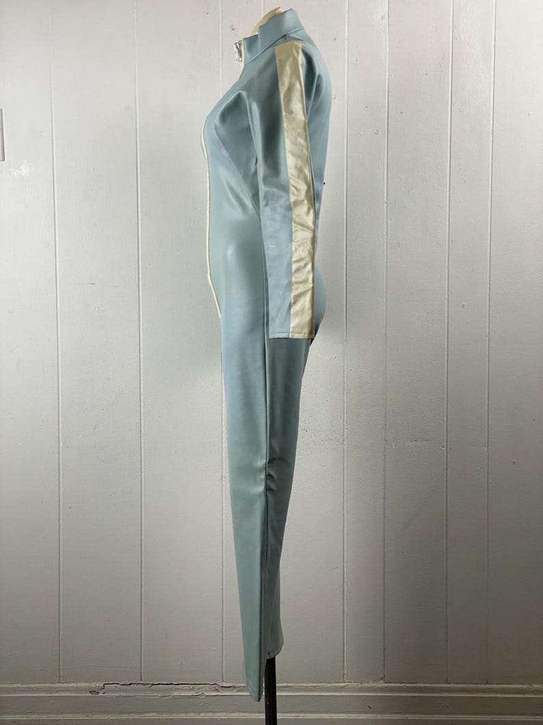K&ouml;nnte beinhalten: Ein hellblauer Jumpsuit mit einem wei&szlig;en Streifen an der Seite des rechten Beins und &Auml;rmels. Der Jumpsuit hat einen Rei&szlig;verschluss an der Vorderseite und einen hohen Kragen.
