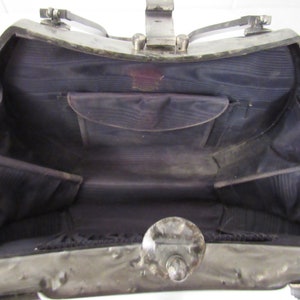 May include: A silver metal handbag with a hinged top and a black fabric interior. The bag has a decorative metal clasp and a small pocket inside.