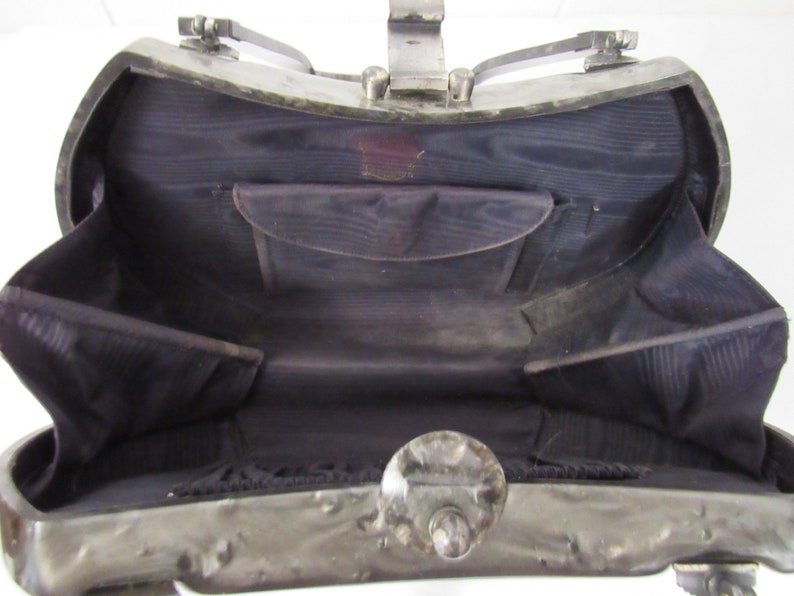 May include: A silver metal handbag with a hinged top and a black fabric interior. The bag has a decorative metal clasp and a small pocket inside.