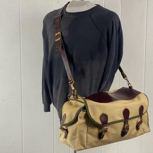Puede incluir: Bolso mensajero de lona beige con solapa y detalles de cuero marrón oscuro. El bolso tiene una correa de cuero marrón y hebillas de latón. El bolso se exhibe en un maniquí con una sudadera gris oscuro.