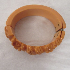 Puede incluir: Un brazalete vintage de pl&aacute;stico de color melocot&oacute;n con un dise&ntilde;o floral tallado. El brazalete tiene bisagras y un cierre de presi&oacute;n.