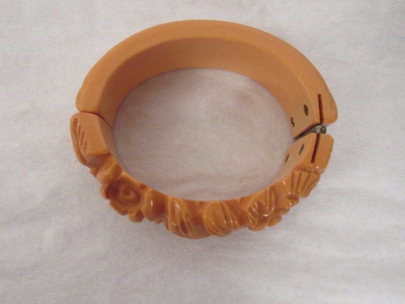 Puede incluir: Un brazalete vintage de pl&aacute;stico de color melocot&oacute;n con un dise&ntilde;o floral tallado. El brazalete tiene bisagras y un cierre de presi&oacute;n.
