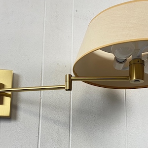 May include: A brass-coloured wall-mounted lamp with a beige lampshade. The lamp has an adjustable arm and a rectangular base. The lampshade is a light beige colour and has a rounded shape. The lamp is mounted on a white wall.