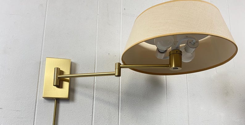 May include: A brass-coloured wall-mounted lamp with a beige lampshade. The lamp has an adjustable arm and a rectangular base. The lampshade is a light beige colour and has a rounded shape. The lamp is mounted on a white wall.