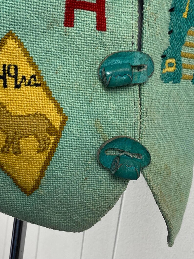 May include: A teal blue vest with a yellow and brown diamond-shaped patch with the text "49ers" embroidered on it. The vest has two teal blue buttons with a decorative design.