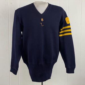 May include: Navy blue V-neck sweater with yellow stripes on the left sleeve and a yellow patch with the number 8. The sweater has a ribbed hem and cuffs, and a small gold-colored pin near the neckline.