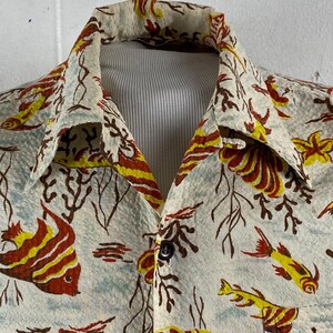 May include: A white button-up shirt with a tropical print of yellow, red, and brown fish and coral. The shirt has a collar and a button closure.
