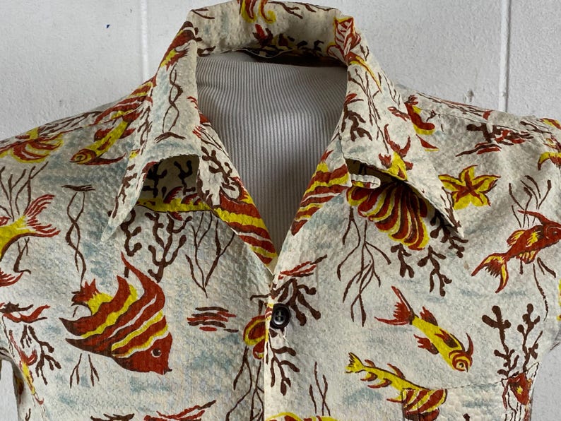 May include: A white button-up shirt with a tropical print of yellow, red, and brown fish and coral. The shirt has a collar and a button closure.