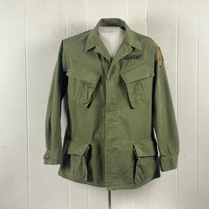 Puede incluir: Chaqueta militar verde oliva con cuatro bolsillos delanteros y cierre de botones. La chaqueta tiene el texto "U.S. ARMY" sobre el bolsillo izquierdo del pecho y un parche rojo en la manga derecha. Prenda militar vintage.
