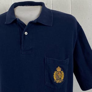 Könnte beinhalten: Marineblaues Poloshirt mit klassischem Kragen und kurzen Ärmeln. Es hat eine Tasche auf der linken Brust mit einem goldfarbenen und mehrfarbigen gestickten Wappen. Das Shirt hat eine Knopfleiste mit drei Knöpfen.