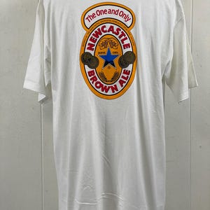 May include: White t-shirt with a large graphic on the back featuring the words "The One and Only Newcastle Brown Ale." The design includes a star, wheat, and the words "Serve Cool."