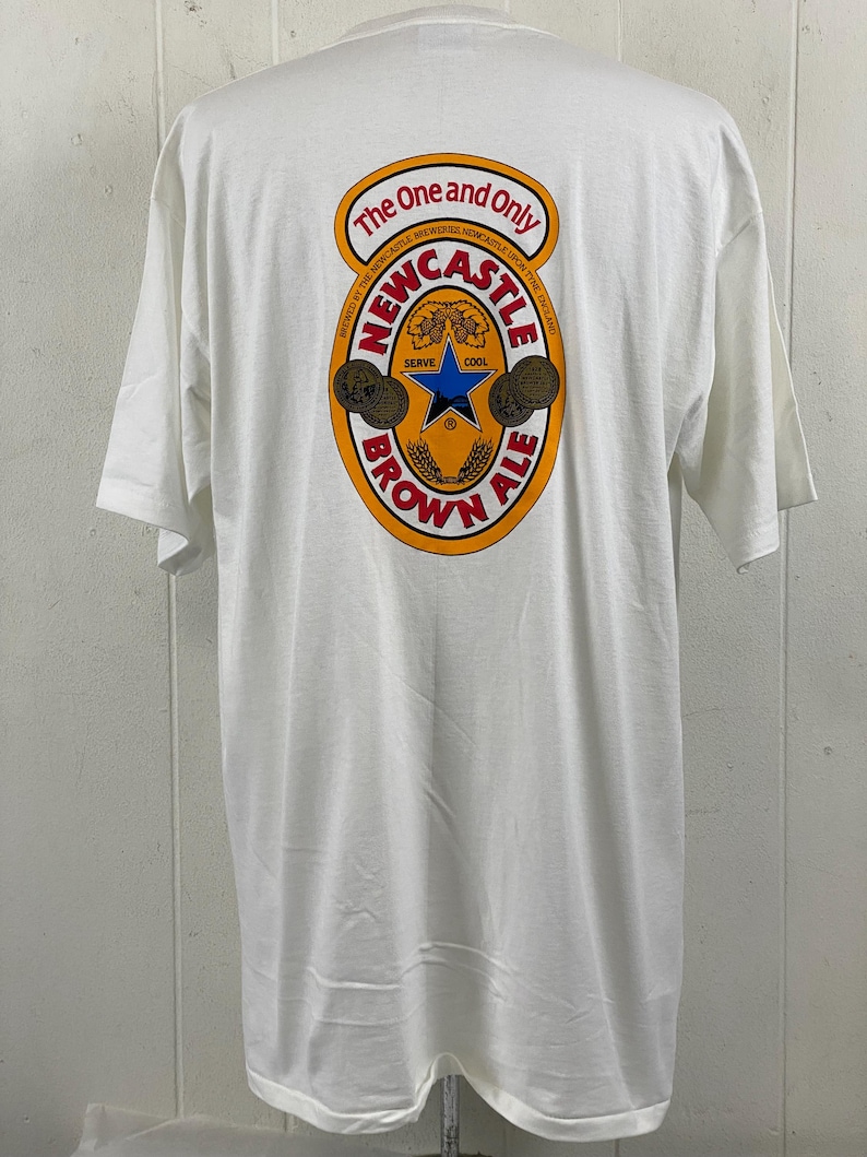 May include: White t-shirt with a large graphic on the back featuring the words "The One and Only Newcastle Brown Ale." The design includes a star, wheat, and the words "Serve Cool."