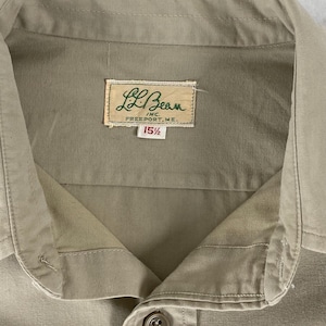 Op de afbeelding: Close-up van een beige button-down shirt met een kraag. Het shirt heeft een label met de tekst "L.L.Bean INC. FREEPORT, ME." en de maat "15 1/2". Het shirt is gemaakt van een zachte, getextureerde stof.