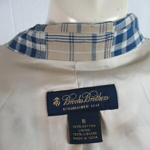 May include: A beige blazer with a blue and tan plaid collar. The label reads "Brooks Brothers Established 1818" and includes the size 16, fabric content, and "Made in India".