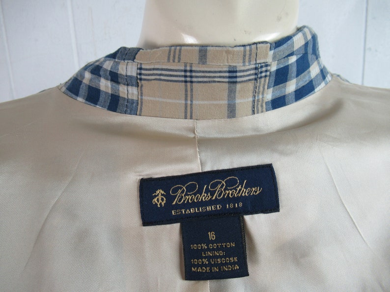 May include: A beige blazer with a blue and tan plaid collar. The label reads "Brooks Brothers Established 1818" and includes the size 16, fabric content, and "Made in India".