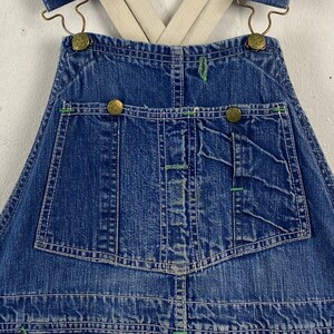 K&ouml;nnte beinhalten: Nahaufnahme einer blauen Jeans-Latzhose mit wei&szlig;en N&auml;hten und gr&uuml;nen Akzenten. Die Latzhose hat messingfarbene Kn&ouml;pfe und verstellbare Hosentr&auml;ger. Das Design umfasst mehrere Taschen und eine klassische Arbeitskleidungs&auml;sthetik.