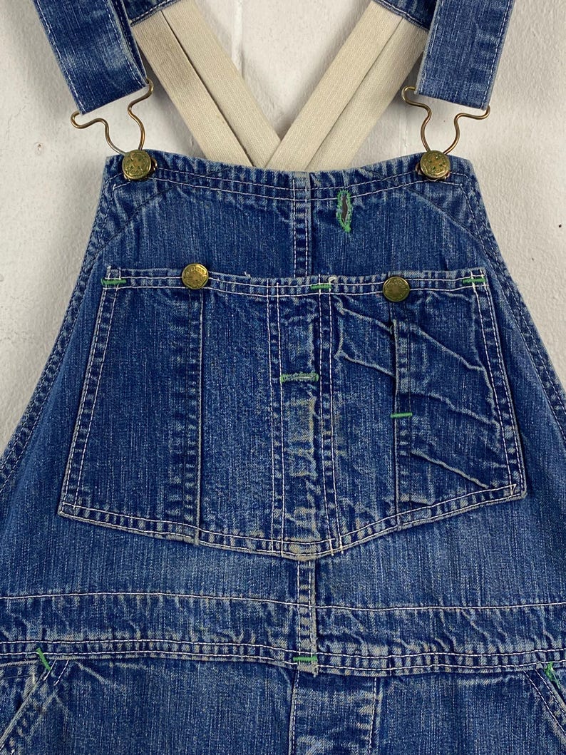 K&ouml;nnte beinhalten: Nahaufnahme einer blauen Jeans-Latzhose mit wei&szlig;en N&auml;hten und gr&uuml;nen Akzenten. Die Latzhose hat messingfarbene Kn&ouml;pfe und verstellbare Hosentr&auml;ger. Das Design umfasst mehrere Taschen und eine klassische Arbeitskleidungs&auml;sthetik.