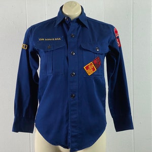May include: A blue long-sleeved button-up shirt with the text "CUB SCOUTS B.S.A." on the left chest pocket. The shirt has two pockets on the front and a variety of patches, including a red and gold patch with a bear cub.