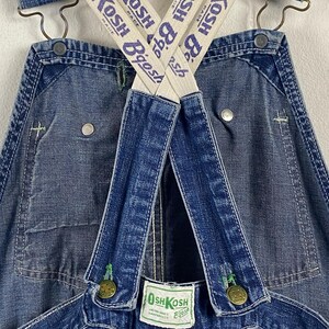 K&ouml;nnte beinhalten: Nahaufnahme einer blauen Jeans-Latzhose mit wei&szlig;-blau gestreiften Tr&auml;gern. Die Tr&auml;ger sind mit "OSH KOSH Bgosh" beschriftet. Die Latzhose hat Metallkn&ouml;pfe und gr&uuml;ne N&auml;hte. Ein kleines Etikett lautet "OSH KOSH Bgosh".