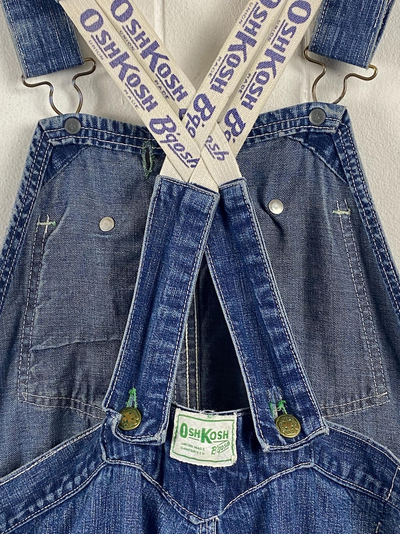 K&ouml;nnte beinhalten: Nahaufnahme einer blauen Jeans-Latzhose mit wei&szlig;-blau gestreiften Tr&auml;gern. Die Tr&auml;ger sind mit "OSH KOSH Bgosh" beschriftet. Die Latzhose hat Metallkn&ouml;pfe und gr&uuml;ne N&auml;hte. Ein kleines Etikett lautet "OSH KOSH Bgosh".