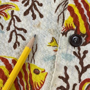 May include: Close-up of a white fabric with a red, yellow, and brown fish print. The fabric has a brown button on the right side.