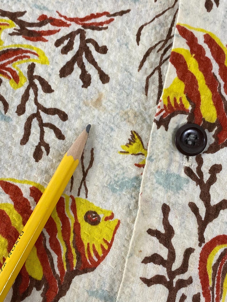 May include: Close-up of a white fabric with a red, yellow, and brown fish print. The fabric has a brown button on the right side.
