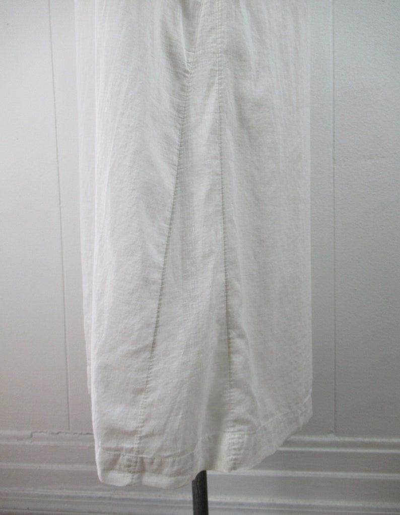 Puede incluir: Un vestido blanco de lino con un dise&ntilde;o sencillo y una falda recta. El vestido tiene una costura en el centro de la espalda y un dobladillo que es ligeramente m&aacute;s largo en la parte posterior.