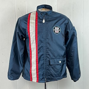 May include: A navy blue zip-up jacket featuring a vertical white stripe flanked by two red stripes on the right side. The left chest displays a circular patch with the text SS CHEVELLE 396. The jacket includes a front pocket with a snap closure.