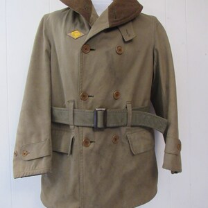 May include: A brown double-breasted military-style coat with a brown fur collar, a belt, and a patch on the left side of the chest. The patch is a yellow diamond with a black symbol inside.