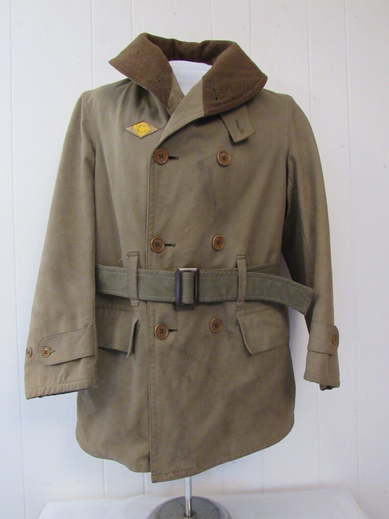 May include: A brown double-breasted military-style coat with a brown fur collar, a belt, and a patch on the left side of the chest. The patch is a yellow diamond with a black symbol inside.